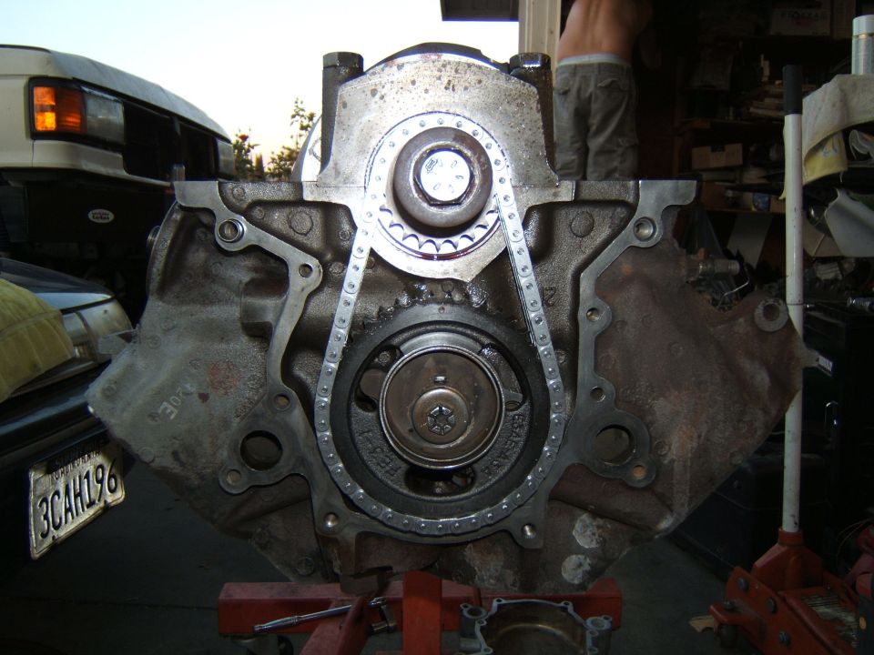 Engine block with timing chain and gears, mounted on an engine stand. A white van is partially visible in the background