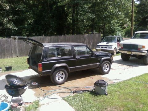 1987 Suzuki Samurai - A black Jeep Cherokee is parked in a driveway with its rear hatch open, alongside other vehicles and outdoor equipment.