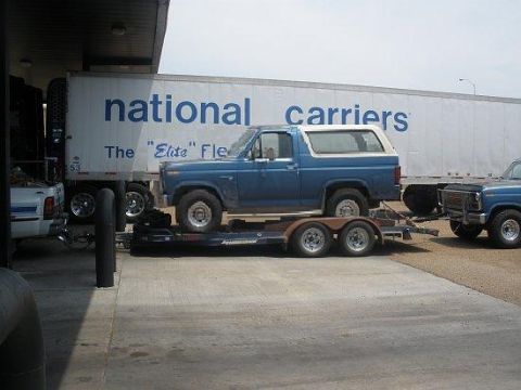 1982 Ford E350 - A blue Ford Bronco is towed on a trailer, with another blue Ford truck visible behind it.