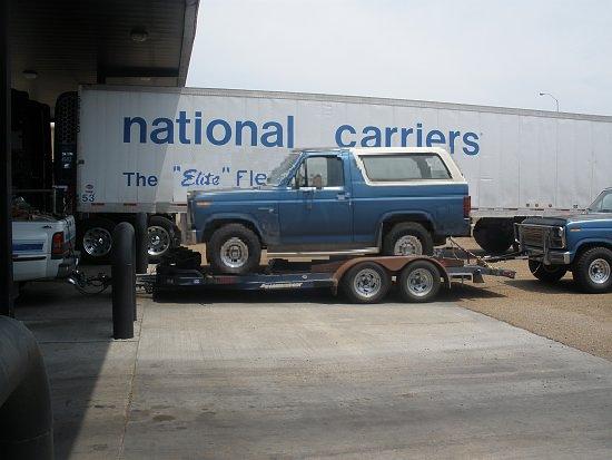Blue Ford Bronco on a trailer, side view. White hardtop. Visible front of another blue Ford truck
