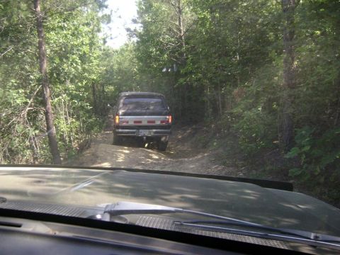 1990 Ford Bronco - A 1990 Ford Bronco navigates a narrow dirt path surrounded by dense trees.