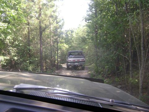 1990 Ford Bronco - A Ford Bronco drives down a wooded dirt road, seen from another vehicle's perspective.
