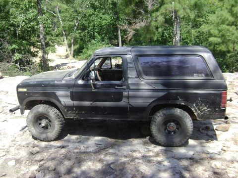1990 Ford Bronco - This black SUV, likely from the early 1980s, is shown parked off-road with prominent side graphics and large tires.