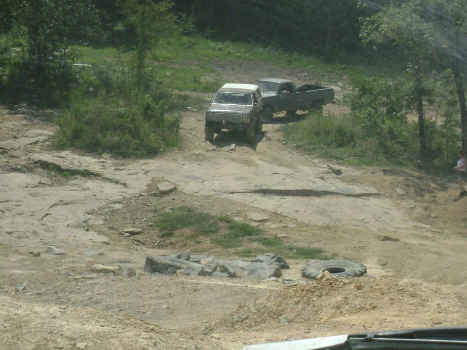 Two trucks navigate a rocky off-road trail with trees and grass on either side