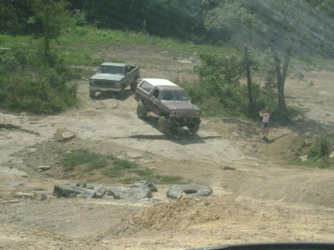 1990 Ford Bronco - Two off-road vehicles navigate a rocky and uneven dirt path surrounded by trees and vegetation.