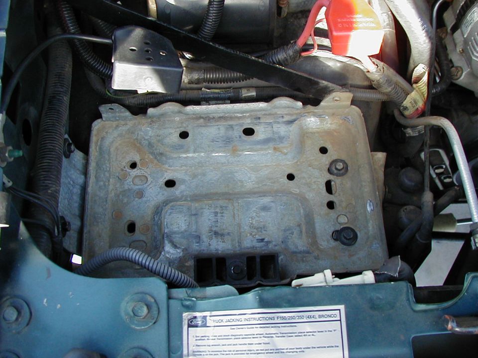 Empty rusty battery tray in engine bay of teal Ford Bronco with red battery cable and jacking instructions decal