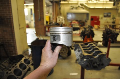 1996 Ford Bronco - A hand holds a single engine piston, with engine blocks and other automotive parts visible in the workshop setting.