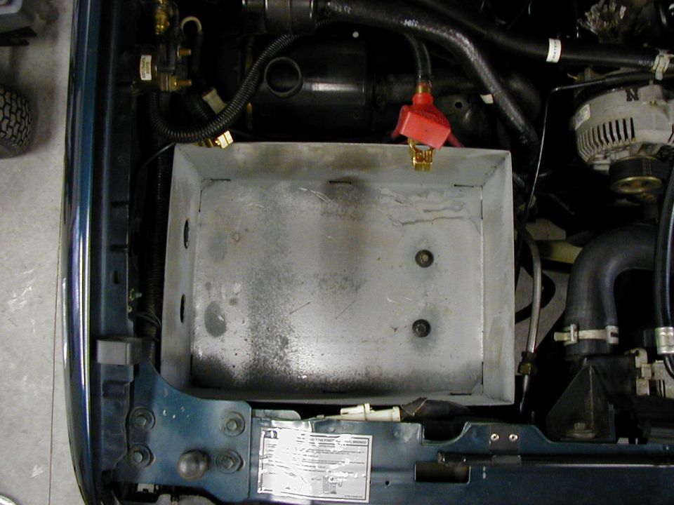 Empty silver battery tray in engine bay with red positive cable terminal resting on edge, battery removed