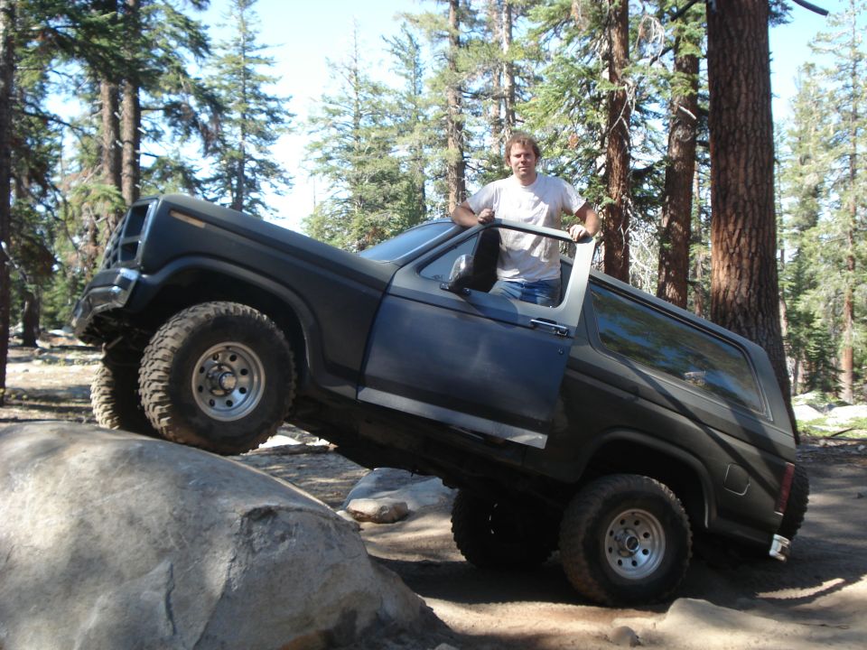 Dark SUV with off-road tires climbing a large rock, driver standing in the open door