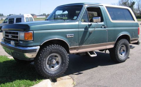 1989 Ford Bronco - This green Ford Bronco has tan trim and aftermarket wheels, and is parked on a curb with a van in the background.