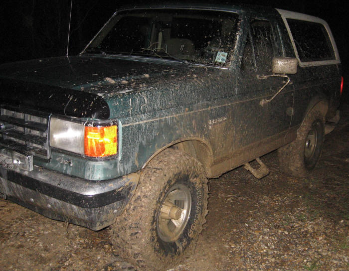 Mud-covered green Ford Bronco, 3/4 front view, with white camper shell