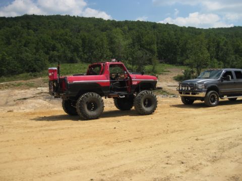 1980 Ford Bronco - Two off-road vehicles parked on a dirt path with wooded hills in the background.