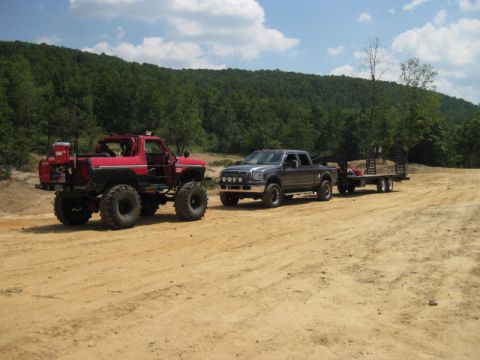 1980 Ford Bronco - Two trucks, a modified red Bronco and a grey pickup towing a trailer, are parked on a dirt road with a wooded hillside in the background.