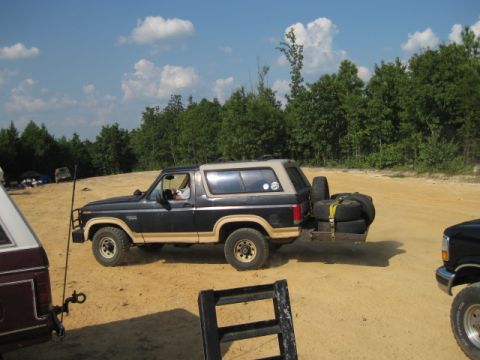 1980 Ford Bronco - A black and tan 1980 Ford Bronco is parked on a dirt lot with spare tires loaded onto a trailer behind it.