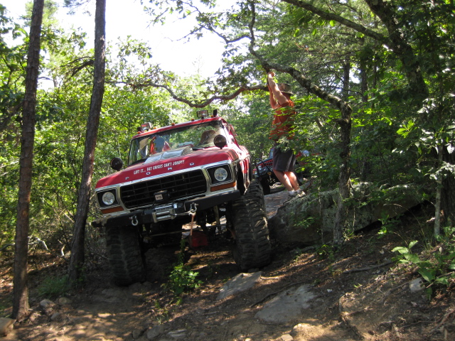 Red off-road truck with large tires climbing a rocky incline, a person hanging from a tree branch above