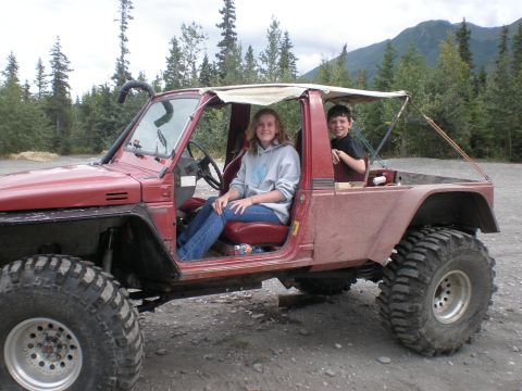 1987 Suzuki Samurai - Two children sit in a modified red off-road vehicle with large tires, parked outdoors.