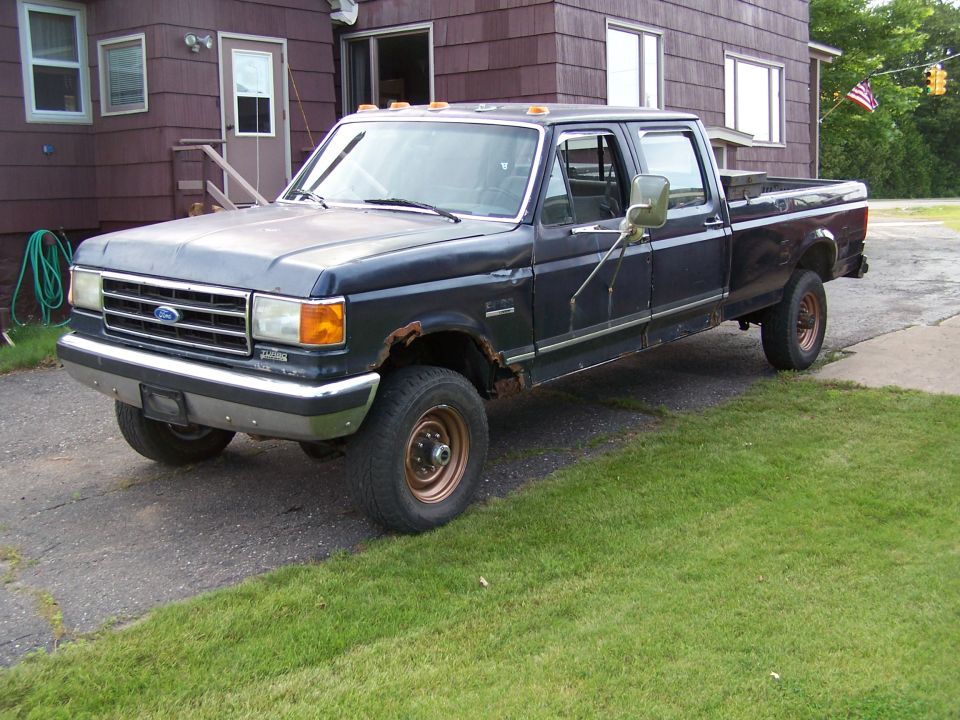 Black Ford crew-cab pickup with rust and faded paint parked on grass by a house