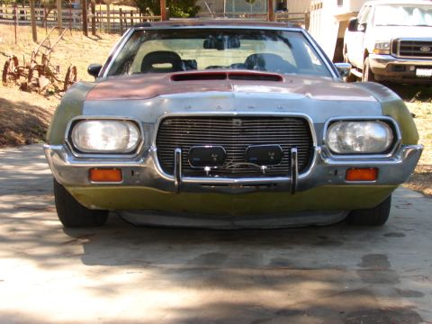 1973 Ford Ranchero - Head-on front view of an olive-green 1970s Ford project car in bare primer and sanded patches across the hood and fenders, parked on a conc…