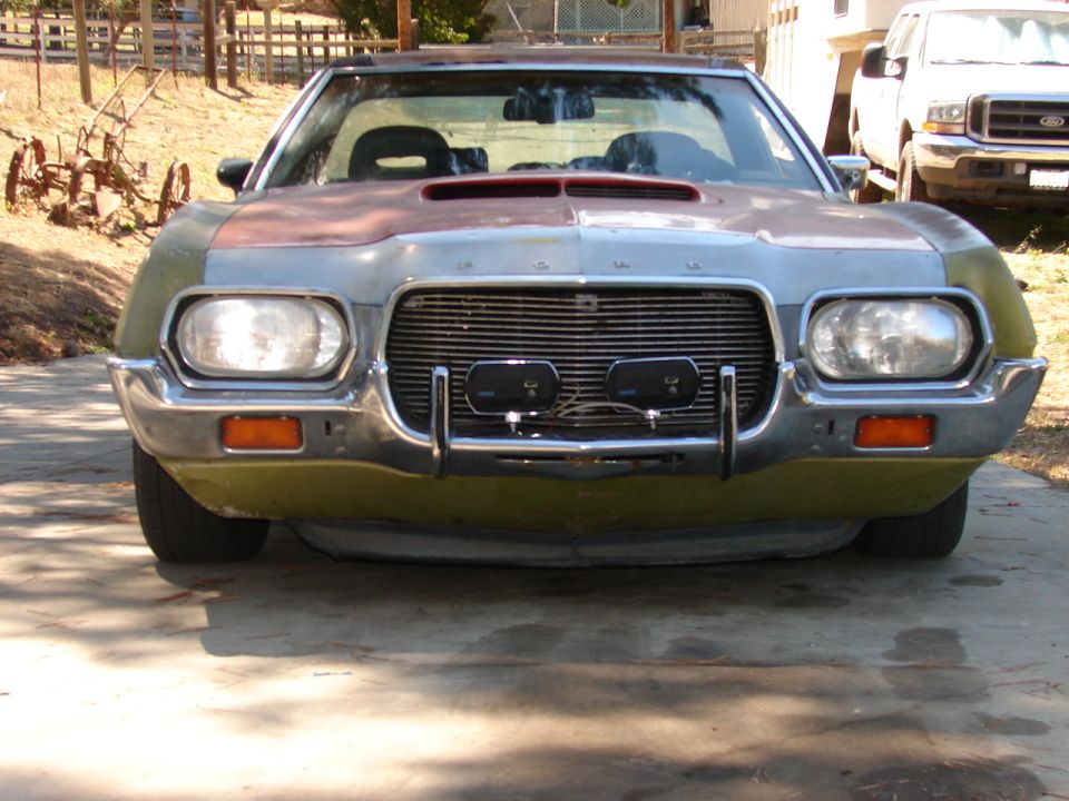 Olive-green 1970s Ford project car front view with hood scoop, chrome bumper and driving lights