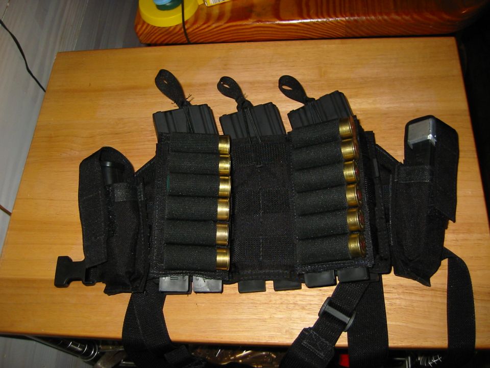 Black tactical chest rig with multiple rifle magazines and shotgun shells, resting on a wooden surface
