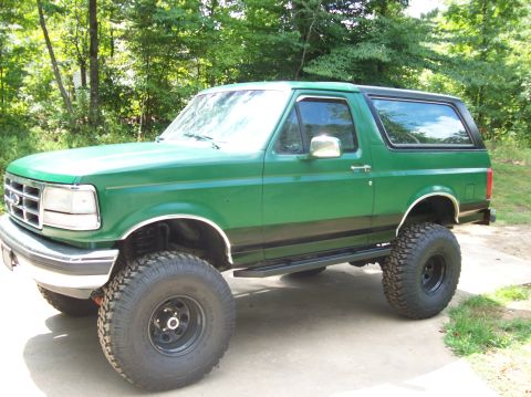 1992 Ford Bronco - This lifted green Ford Bronco is shown from the front three-quarter view, parked on a paved surface with trees behind it.