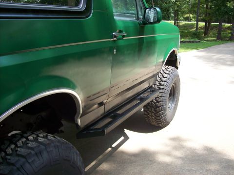 1992 Ford Bronco - This green SUV features large off-road tires and aftermarket side steps, parked on a paved surface with trees in the background.