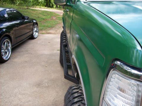 1992 Ford Bronco - A green 1992 Ford Bronco is parked next to a black car, showing its side steps and large tires.