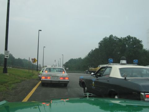 1991 Ford Crown Victoria - A dark police car with blue lights and a white roof is parked on the side of the road, with another police car ahead.