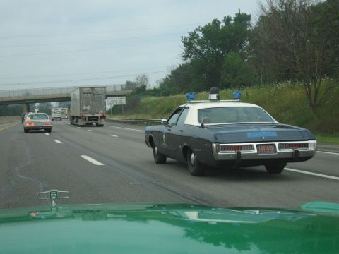 1991 Ford Crown Victoria - A police car with a light bar and blue lights drives on the highway. Another vehicle and a truck are visible ahead.