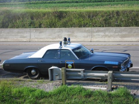 1991 Ford Crown Victoria - Side view of a 1977 Dodge Monaco police car with a light bar on the roof, parked by a highway.