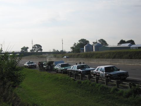 1991 Ford Crown Victoria - A line of police cars and a tow truck are pulled over on the highway, with some activity around the tow truck.