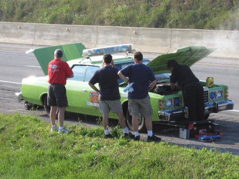 1991 Ford Crown Victoria - Four men inspect a lime green sedan with its hood open and emergency lights on the roof, stopped on the side of a highway.
