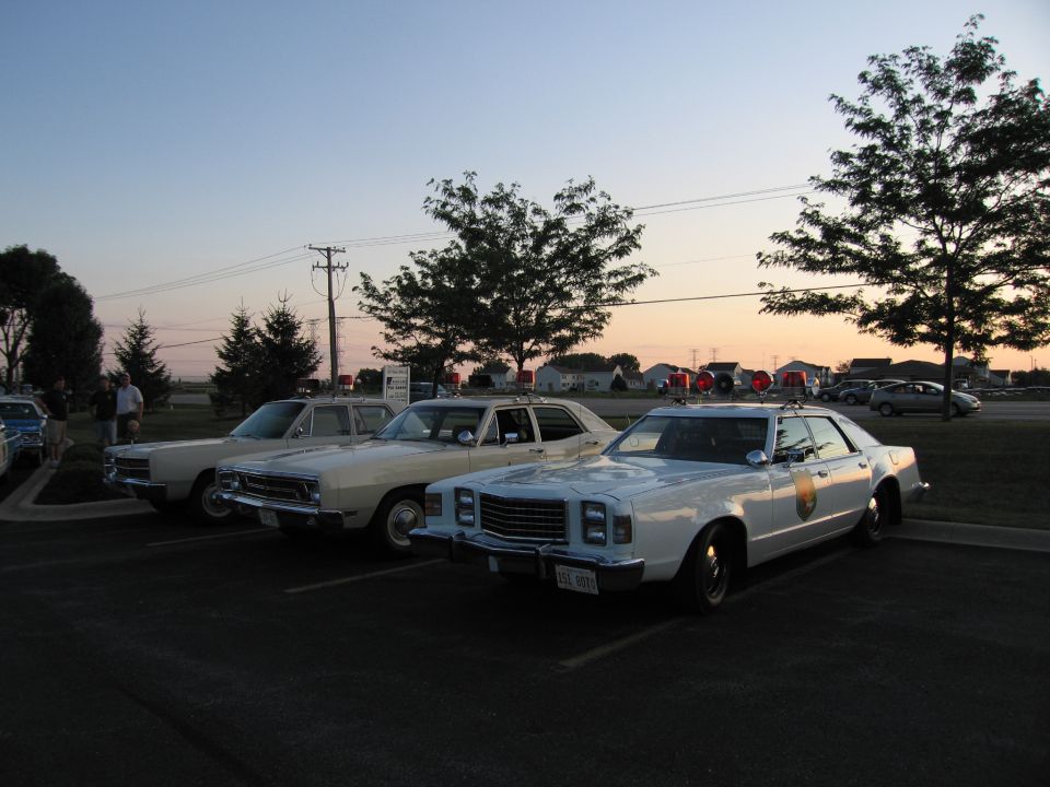 White vintage police sedan with door emblem parked beside other cars at dusk