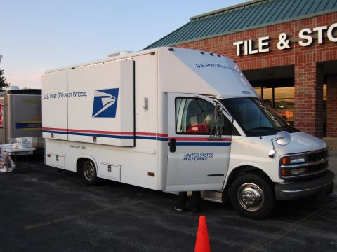 1991 Ford Crown Victoria - A white United States Post Office truck is parked in front of a brick building with a sign that reads 'TILE & STORE'.