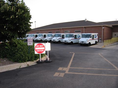 1991 Ford Crown Victoria - Multiple white USPS delivery trucks are parked in a lot outside a brick building.