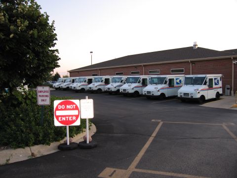 1991 Ford Crown Victoria - A fleet of USPS delivery trucks are parked in a lot outside a brick building, with 'Do Not Enter' and 'Employee Parking Only' signs visible.