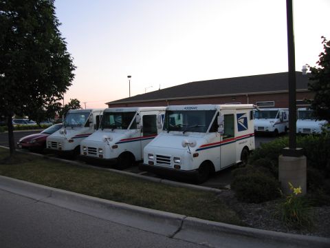 1991 Ford Crown Victoria - Multiple USPS delivery trucks are parked in a lot outside a building.
