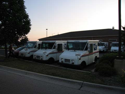 1991 Ford Crown Victoria - A line of white postal service trucks are parked outside a building at dusk.