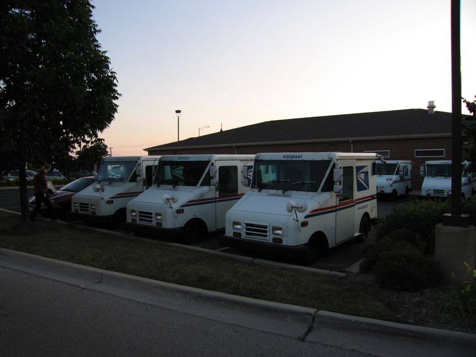 USPS mail trucks parked at a curb with a person walking by at sunset
