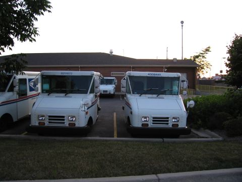 1991 Ford Crown Victoria - Multiple white USPS delivery trucks are parked in a lot outside a brick building.