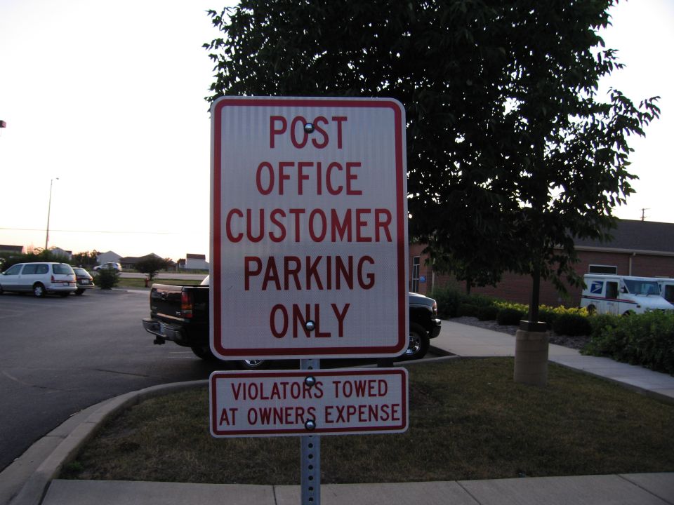 Red and white "Post Office Customer Parking Only, Violators Towed" sign in a parking lot