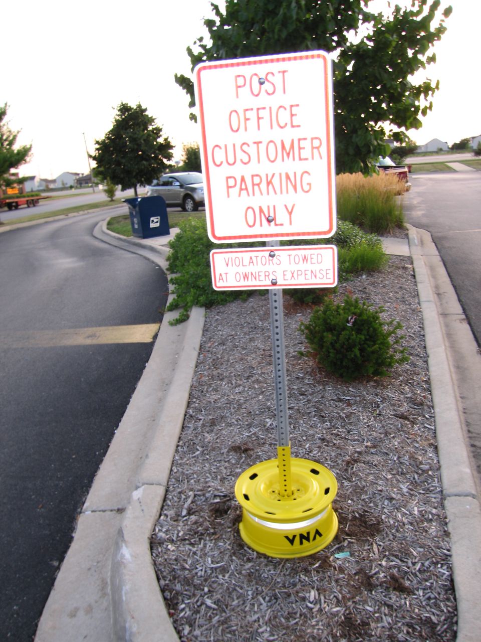 Post office parking sign on a post in a yellow rim base in a curbside mulch median