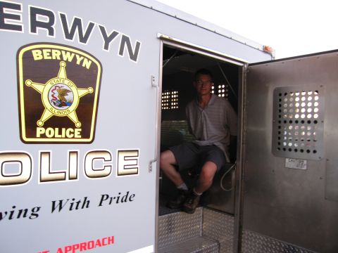 1991 Ford Crown Victoria - A man is seated in the back of a Berwyn Police van, which has the Illinois state police emblem visible on its side.