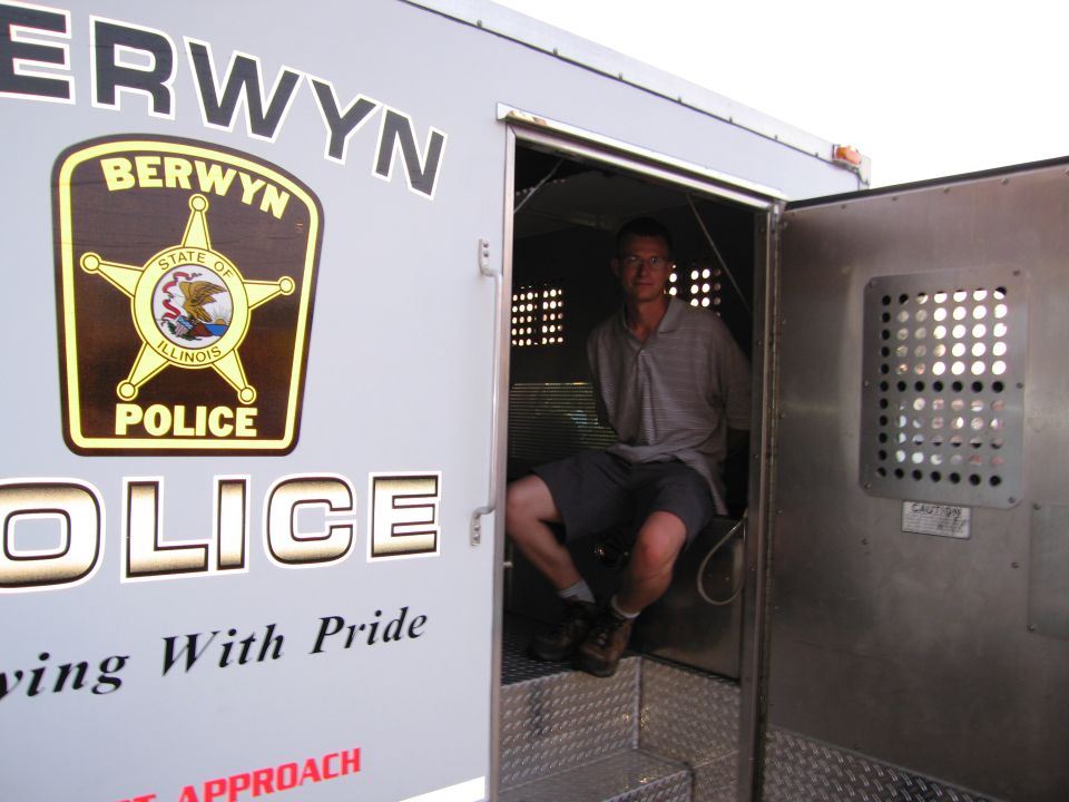 Man seated in open rear cage of a white Berwyn Police transport van