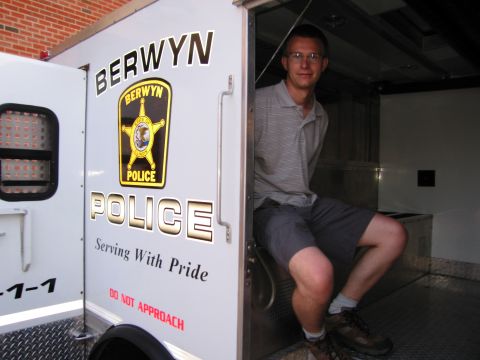 1991 Ford Crown Victoria - A man sits in the back of a Berwyn Police vehicle, which has "Serving With Pride" written on its side.
