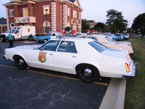 1991 Ford Crown Victoria - A white police car is parked in a lot with other vehicles and a brick building in the background.