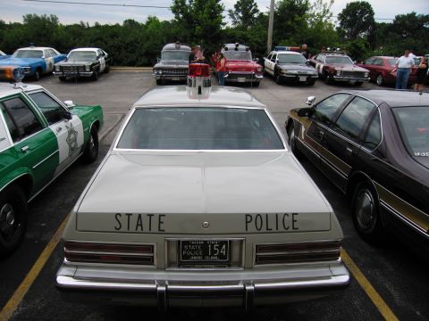 1991 Ford Crown Victoria - Several police cars are parked in a lot, including a light-colored car with 'STATE POLICE' visible on its rear.