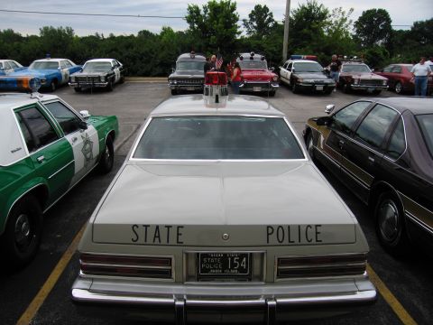 1991 Ford Crown Victoria - A collection of marked police vehicles are gathered together in a parking lot under an overcast sky.