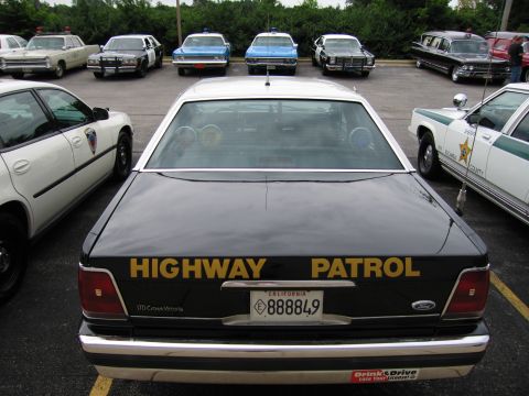 1991 Ford Crown Victoria - A lineup of police vehicles, including a black Ford Crown Victoria marked 'HIGHWAY PATROL', is parked in a lot.