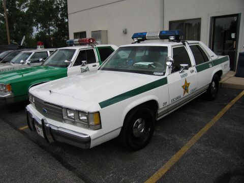 1991 Ford Crown Victoria - Two Sheriff's vehicles, a white Ford Crown Victoria and a green car, are parked side-by-side.